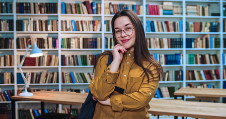 Portrait of playful friendly brunette student with stylish black backpack collecting information from different books in library.