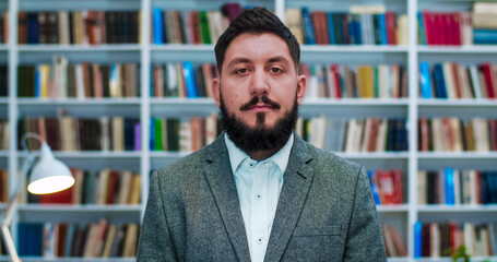 Young Caucasian businessman standing in cabinet with books shelves and looking straight to camera. Portrait of man with beard in public library or book store. Male professor.