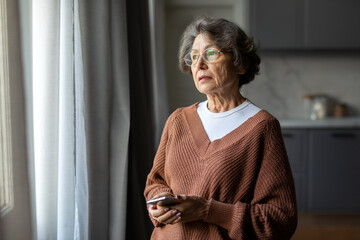Worried depressed senior woman stand by window look away troubled, holding cellphone in hands, lady...