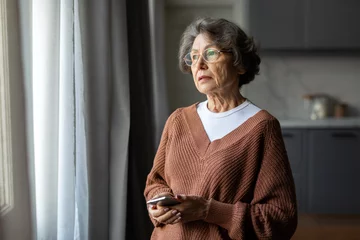 Fotobehang Worried depressed senior woman stand by window look away troubled, holding cellphone in hands, lady lost in sad heavy thoughts © Home-stock