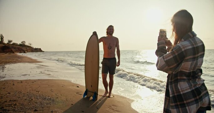 A blonde girl in a plaid shirt takes a photo of her Happy boyfriend with a naked torso in gray shorts, who holds a surfboard in his hands and stands on the sandy seashore in the morning at Sunrise