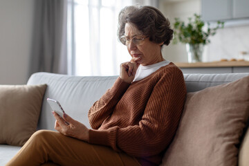 Serious senior woman using smartphone and frowning reading bad news online, sitting on couch at...
