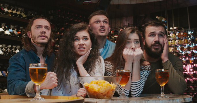 Young football male and female fans watching game together in evening in pub and cheering up for their favorite team. Caucasian friends are upset and dissapointed of soccer loose in bar.