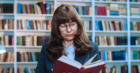 Close-up portrait of brunette female professor in glasses working and reading book in library. Bookcase in background.