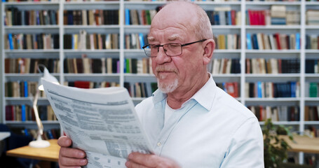 Portrait of old Caucasian bald man in glasses reading newspaper in library and looking to camera....