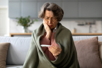 Sick senior caucasian woman checking her temperature, sitting wrapped in blanket on sofa, elderly...