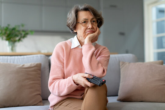 Bored elderly lady sitting on the couch in living room, pointing at tv remote to switch channels, unhappy with wifi issues or television program