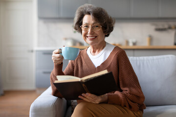 Happy elderly senior woman drinking hot tea and reading favorite paper book, enjoying cozy weekend,...