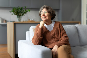Dreamy senior woman in comfy outwear and eyeglasses resting on couch in living room, looking aside...