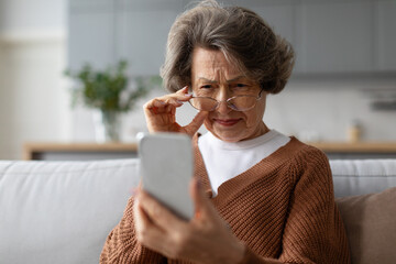 Senior european woman squinting eyes looking at phone screen, wearing eyeglasses sitting on couch at home. Bad vision health problems - obrazy, fototapety, plakaty