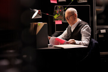 Elderly private detective sitting at desk table in dark room, working overtime at criminal case....