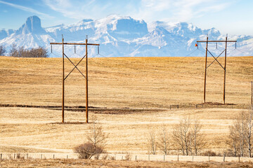 Pair of large wooden power poles with hanging electrical wires overlooking a hilltop and the distant Canadian Rocky Mountains near Cochrane Alberta Canada.