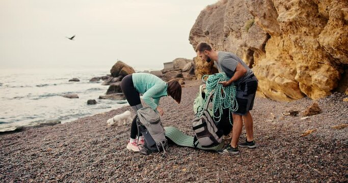 A group of rock climbers, two girls and a brunette guy came to the rocky seashore and lay out their backpacks in order to put on climbing equipment near the yellow rocks on the sea during the day