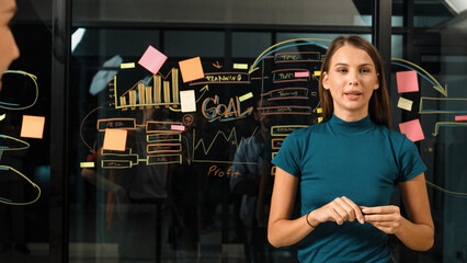 Obraz na płótnie Canvas Skilled manager standing at glass wall while looking at camera. Professional caucasian businesswoman or female leader present marketing strategy while using sticky notes and mind mapping. Tracery