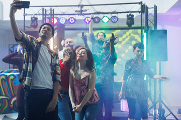 Man clubber taking selfie with women while partying at nightclub discotheque. Young friends posing for mobile phone photo while dancing and having fun on club dancefloor illuminated with lights