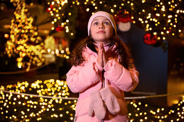 Little girl with cupped hands, makes cherished wish for Christmas, standing at fairground, enjoying...