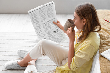 Beautiful young woman with cup of tea reading newspaper in bedroom at home