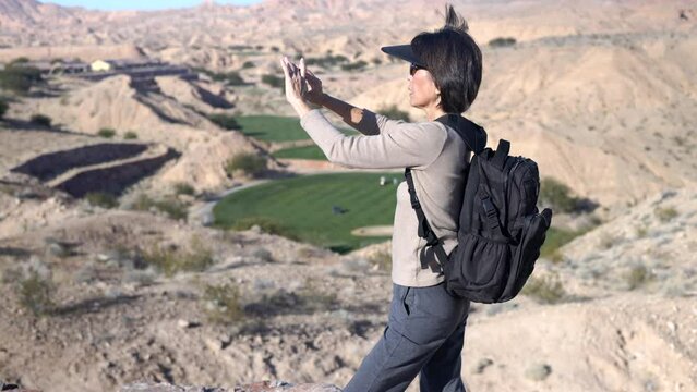 Woman wearing backpack and visor walking in desert stops to take photo of distant view with golf fairway