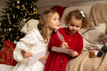 Two little girls write letters to Santa Claus near the Christmas tree indoors.