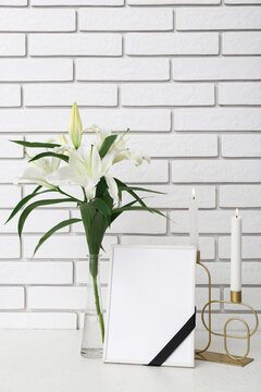 Blank funeral frame, burning candles and vase with lily flowers on table near light brick wall
