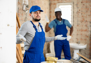 Caucasian man standing in construction site and making confused gesture. His foreman, african-american man, standing in background and shouting.