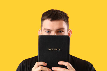 Young priest with Holy Bible on yellow background, closeup - obrazy, fototapety, plakaty