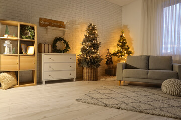 Interior of festive living room with grey sofa, Christmas trees and glowing lights at evening