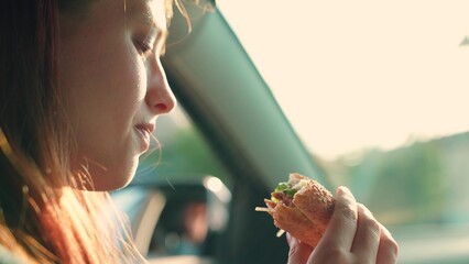 Young woman takes bite of burger while traveling in car. Young woman indulges in quick cuisine....