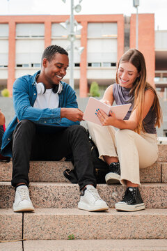 Vertical. Two multiracial teenage studens learning together at university campus. African american schoolboy talking with a school girl about the homeworks. Young friends studying at highschool stairs