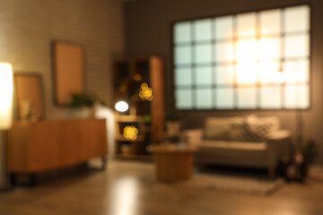 Blurred view of modern living room with grey sofa, coffee table, wooden cabinet and glowing lamps at evening