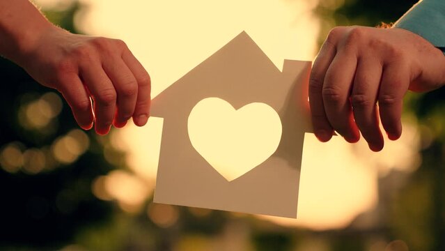 Symbol of home, happiness. Concept of building house for family, child. Real estate insurance. Paper house in hands at sunset in park, sun is shining through window, family dreams of buying house.