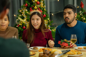 Group of Asian friends enjoying Christmas party for new year celebrate. Friends having fun together with dinner at home. men and women opening box gift and present together at home with Christmas tree