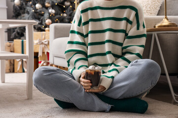Obraz premium Woman holding glass of hot chocolate with marshmallows in festive living room, closeup