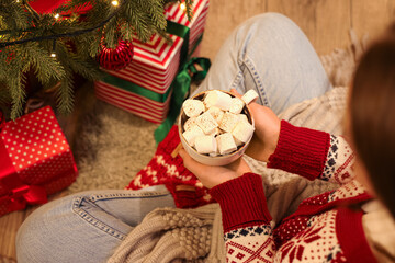 Woman with cup of hot chocolate sitting on floor near Christmas tree, closeup