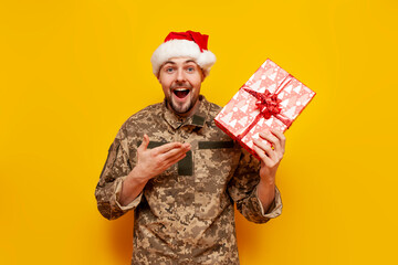 Ukrainian soldier in pixelated uniform and santa claus hat holding gift box on yellow isolated background