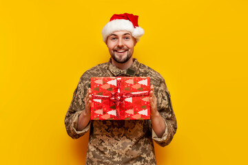 Ukrainian soldier in pixelated uniform and santa claus hat holding gift box on yellow isolated...