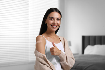 Woman with sticking plaster on arm after vaccination showing thumbs up in bedroom