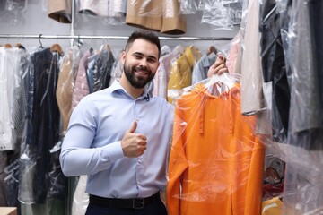 Dry-cleaning service. Happy worker holding hanger with hoodie in plastic bag and showing thumb up...