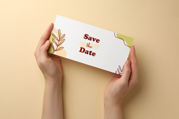 Woman holding beautiful card with Save the Date phrase on beige background, top view