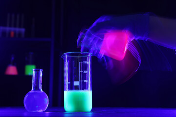 Scientist working with laboratory glassware of luminous liquids at table against dark background,...