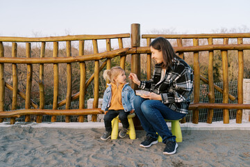 Mom feeds a little girl from a spoon while sitting on chairs near a wooden fence