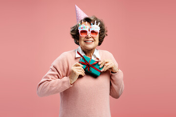 Portrait attractive smiling senior woman, happy 70 years old grandmother wearing funny eyeglasses holding gift box celebration birthday isolated on pink background. Holiday party concept  - obrazy, fototapety, plakaty