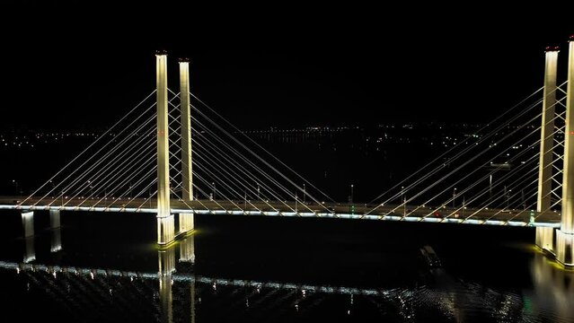 A cable-stayed highway bridge with tall concrete pylons illuminated by architectural lighting at night. General plan, the camera flies forward and circles through the bridge supports