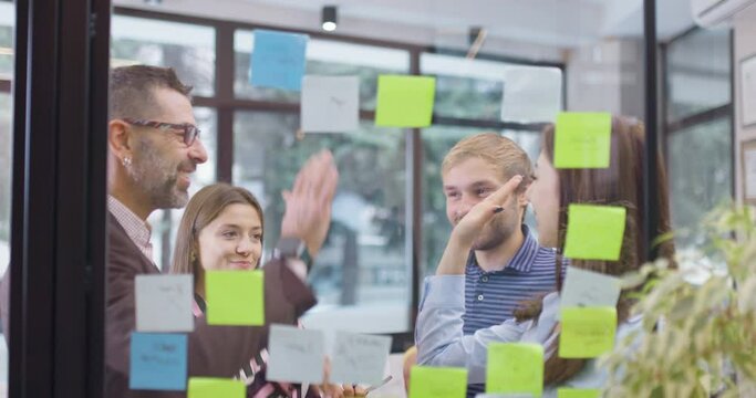 Work colleagues discussing startup project ideas and giving high five. Caucasian business team at glass wall with sticky notes. Scrum with colorful stickers. Vertical movement.