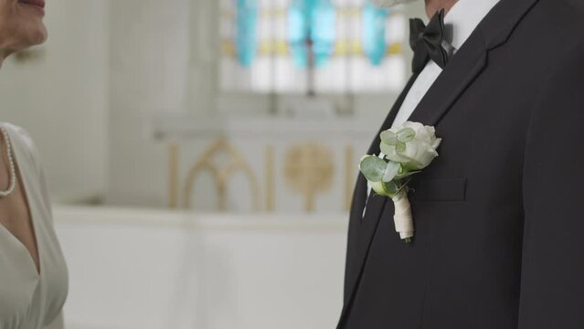 Cropped shot of Caucasian mid-aged bride in elegant white wedding dress and pearl necklace adjusting white rose buttonhole on her groom suit, having wedding ceremony in church