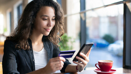 Young woman holding credit card and using mobile phone in cafe. Online shopping concept - obrazy, fototapety, plakaty