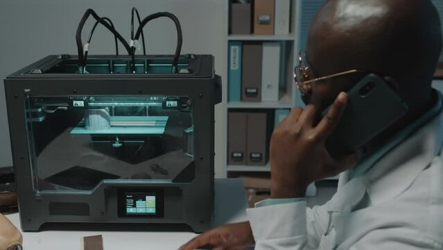 African American lab scientist talking over phone while sitting and testing new 3D printing machine