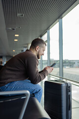 portrait of cheerful man relaxing at airport with bag and mobile phone, guy waiting for flight...
