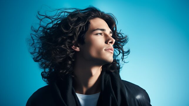 young handsome male model with long black curly hair looking away isolated on blue background with copy space, stylish young man portrait on blue.
