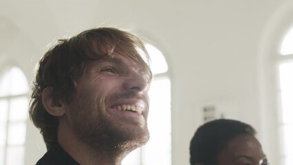 Low angle medium closeup with slowmo of handsome young Caucasian best man crying tears of joy, smiling and clapping while sitting at bench with his Black wife during wedding ceremony in church